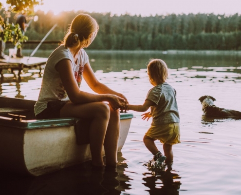 Frau mit Kind sitzen am See und schauen auf ihren Hund im Wasser