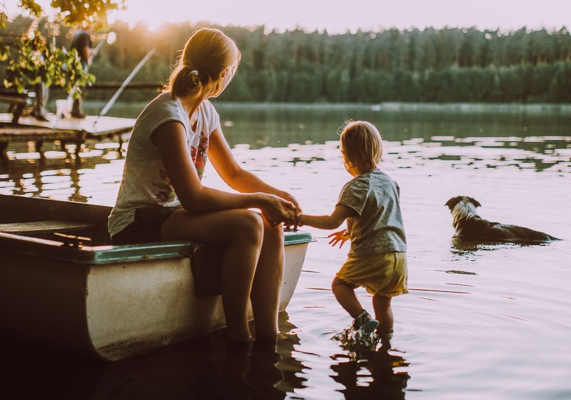 Frau mit Kind sitzen am See und schauen auf ihren Hund im Wasser